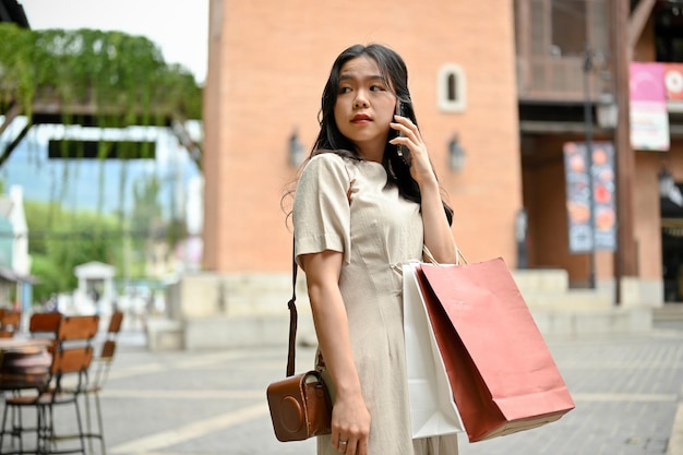 Uma linda mulher asiática está ao telefone com sua amiga enquanto faz compras sozinha na praça da cidade