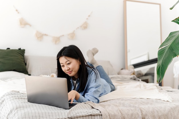 Uma linda mulher asiática está deitada na cama e trabalhando usando um laptop em casa Uma jovem trabalha em casa em quarentena distanciamento social auto-isolamento