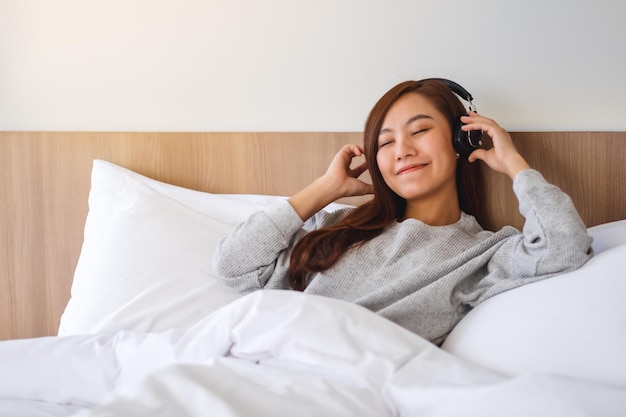 Uma linda mulher asiática gosta de ouvir música com fone de ouvido enquanto está deitada em uma cama branca e aconchegante em casa