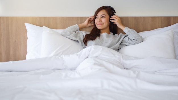 Uma linda mulher asiática gosta de ouvir música com fone de ouvido enquanto está deitada em uma cama branca e aconchegante em casa