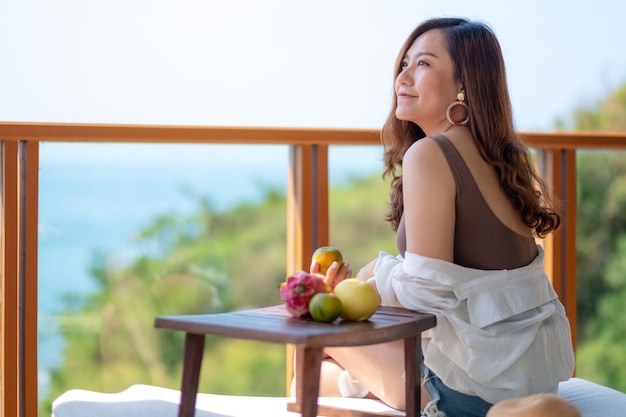 Uma linda mulher asiática segurando frutas enquanto se senta na varanda com vista para o mar.