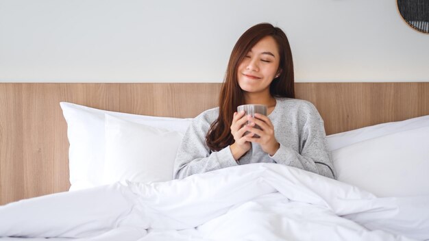 Uma linda mulher asiática tomando café quente enquanto estava deitada em uma cama branca e aconchegante em casa pela manhã