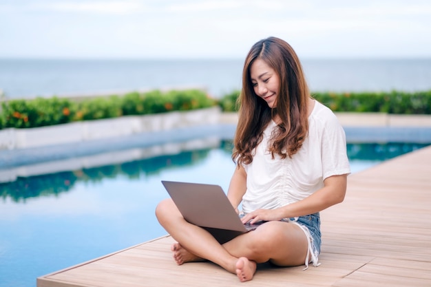 Uma linda mulher asiática usando e digitando no laptop enquanto está sentado à beira da piscina