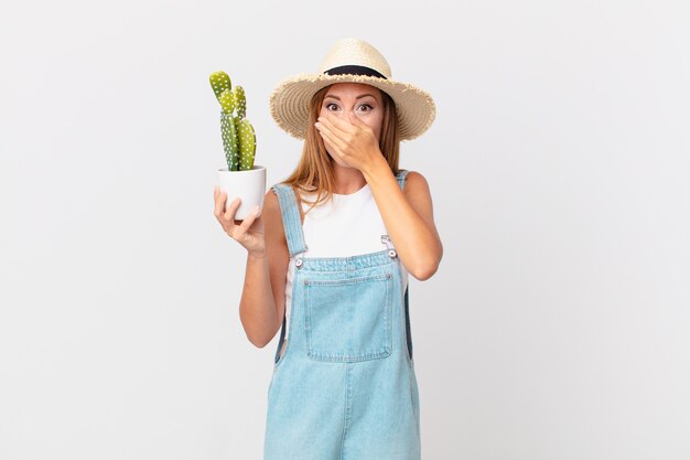 Uma linda mulher cobrindo a boca com as mãos em choque e segurando uma planta decorativa de cacto