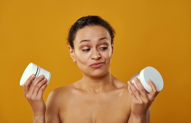 Uma linda mulher com o rosto enrugado ao escolher potes de cremes isolados em fundo amarelo