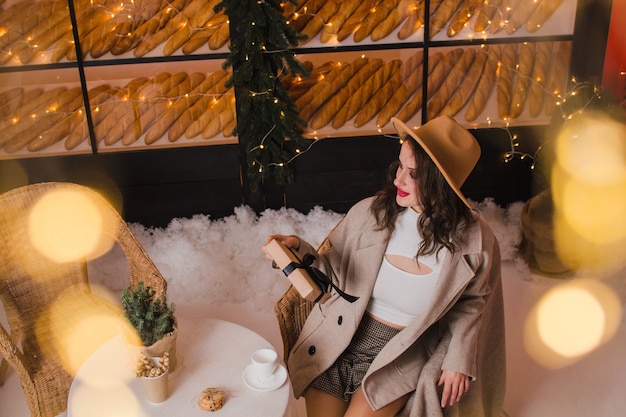 Uma linda mulher com um casaco e um chapéu sentada em uma cafeteria com um presente nas mãos conceito de natal