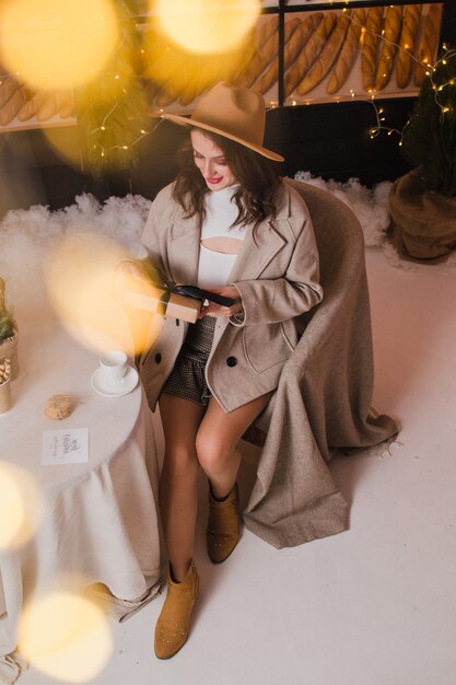 Uma linda mulher com um casaco e um chapéu sentada em uma cafeteria com um presente nas mãos Conceito de Natal