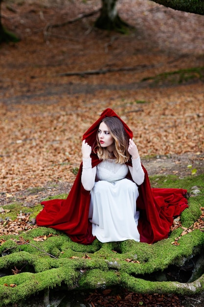 Uma linda mulher com um longo manto vermelho e vestido branco está agachada sobre as raízes de uma árvore