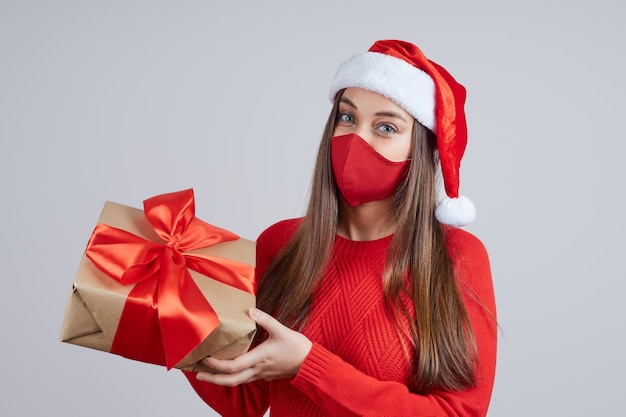 Uma linda mulher com uma máscara protetora e um chapéu de Papai Noel segurando um presente com um laço vermelho