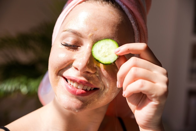 Uma linda mulher com uma toalha rosa na cabeça e um pedaço de pepino no olho