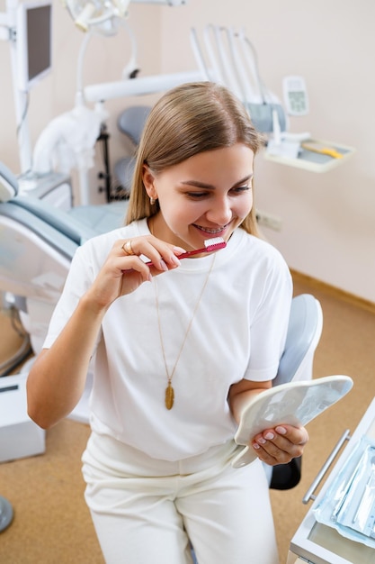 Uma linda mulher de camiseta branca senta-se em um consultório odontológico, olha no espelho, sorri e segura uma escova de dentes nas mãos. Garota com aparelho mostra cuidados bucais. Odontologia, tratamento odontológico