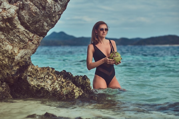 Uma linda mulher de maiô, segura coco com um tubo, de pé em uma praia perto de grandes pedras de recife, goza de férias em uma bela ilha.