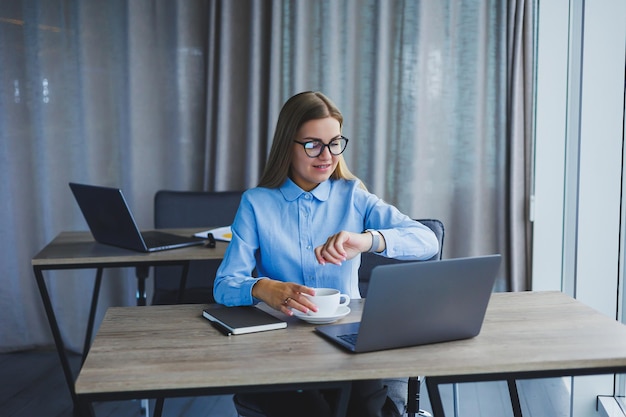 Uma linda mulher de negócios em uma camisa e óculos senta-se com um laptop no escritório à mesa Gerente de mulher de óculos no local de trabalho no escritório moderno Dia de trabalho no computador