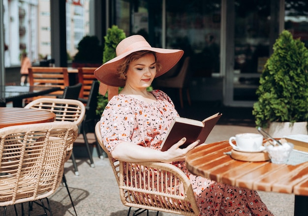 Uma linda mulher de vestido e chapéu está lendo um livro em um café de rua mulher elegante em um vestido de verão