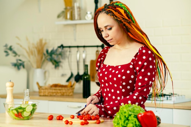 Uma linda mulher de vestido vermelho e dreadlocks está cortando um tomate cereja para uma salada em uma cozinha moderna Vitaminas nutrição dietética e esportiva Comida vegetariana