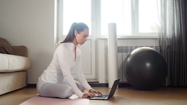 Uma linda mulher em um agasalho de treino branco surfa na internet com um laptop moderno para encontrar novos exercícios no tapete perto do sofá em uma sala iluminada