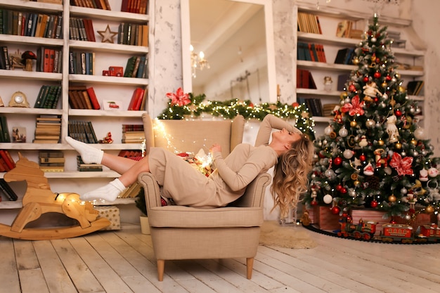 Uma linda mulher em um vestido de casa e meias quentes relaxa em frente à lareira e árvore de Natal