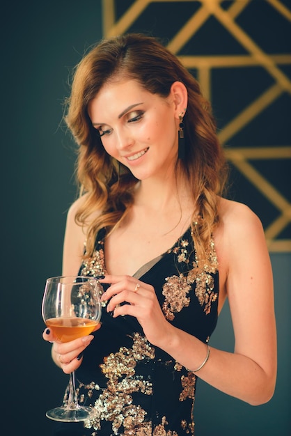 Uma linda mulher em um vestido preto manto no interior escuro bebe conhaque de vinho. Efeito de foto retrô