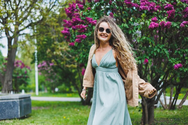 Uma linda mulher encaracolada em um vestido de verão corre e se alegra por estar usando óculos escuros contra o fundo de um arbusto lilás roxo