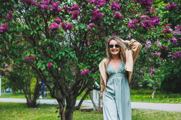 Uma linda mulher encaracolada em um vestido de verão corre e se alegra por estar usando óculos escuros Contra o fundo de um arbusto lilás roxo