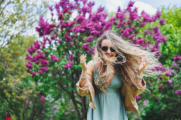 Uma linda mulher encaracolada em um vestido de verão corre e se alegra por estar usando óculos escuros contra o fundo de um arbusto lilás roxo