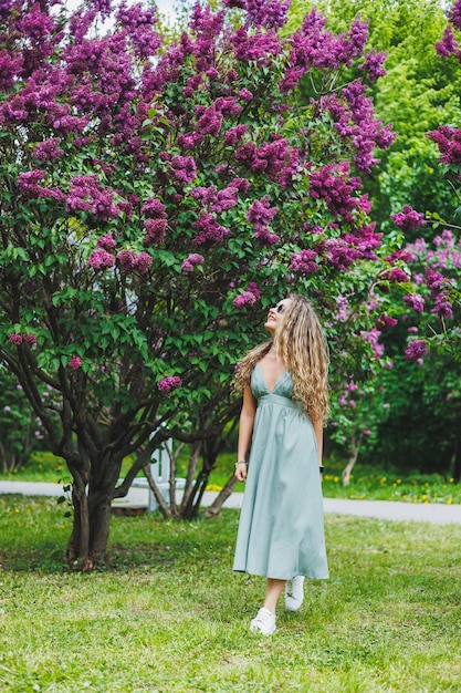 Uma linda mulher encaracolada em um vestido de verão corre e se alegra por estar usando óculos escuros Contra o fundo de um arbusto lilás roxo