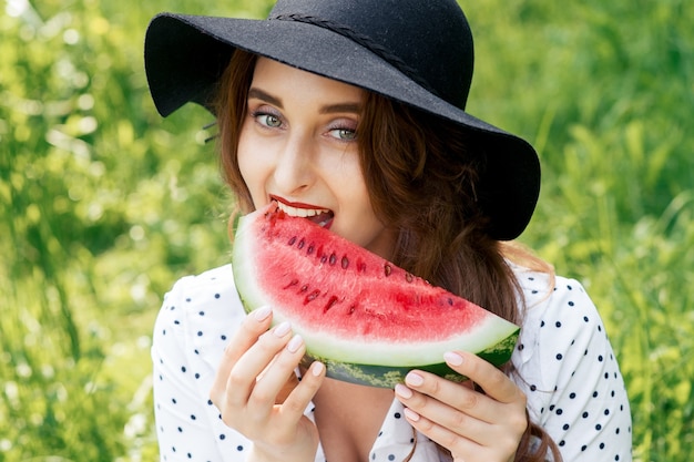 Uma linda mulher está comendo uma fatia de melancia fresca com chapéu preto sobre fundo de grama verde.