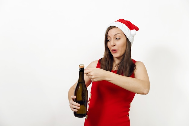 Uma linda mulher feliz caucasiana jovem engraçada com um sorriso encantador no vestido vermelho e chapéu de Natal, tentando abrir a garrafa de champanhe no fundo branco. Santa menina isolada. Conceito de feriado de ano novo