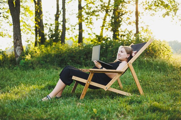 Uma linda mulher grávida está sentada em um parque e trabalhando remotamente em um laptop trabalho on-line em licença de maternidade mulher grávido moderna