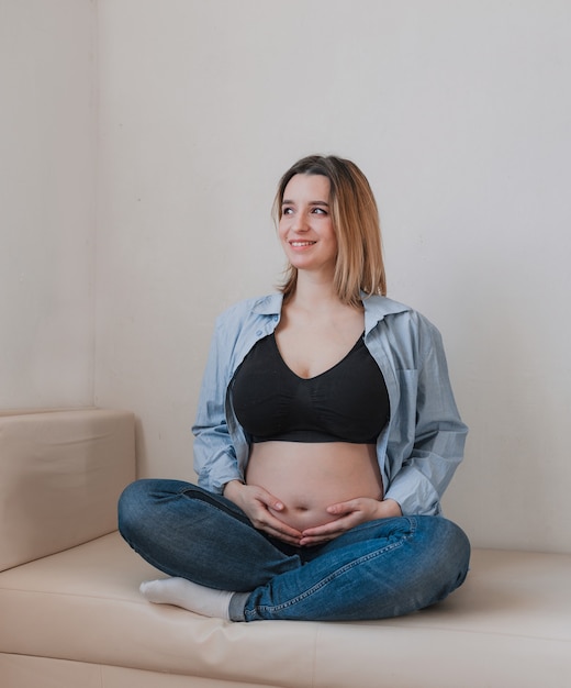 Uma linda mulher grávida está sentada no sofá sorrindo e olhando pela janela. Felicidade feminina