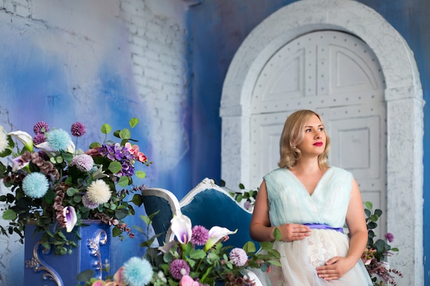 Uma linda mulher grávida senta-se entre flores em uma foto em uma parede azul em estilo loft