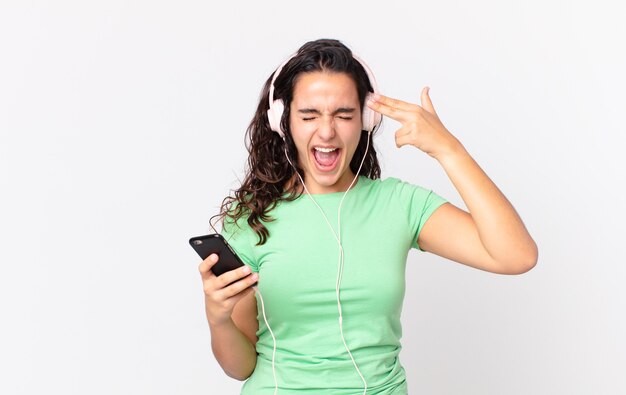 Uma linda mulher hispânica parecendo infeliz e estressada, gesto suicida fazendo sinal de arma com fones de ouvido e um smartphone