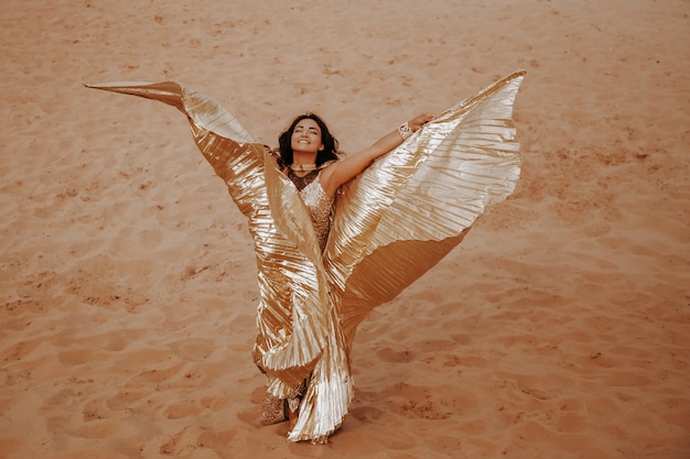 Uma linda mulher loira de dourado com vestido brilhante com asas, terno está dançando uma dança oriental, oriental no deserto na areia. dançarina linda mulher exótica em um traje para dança do ventre.