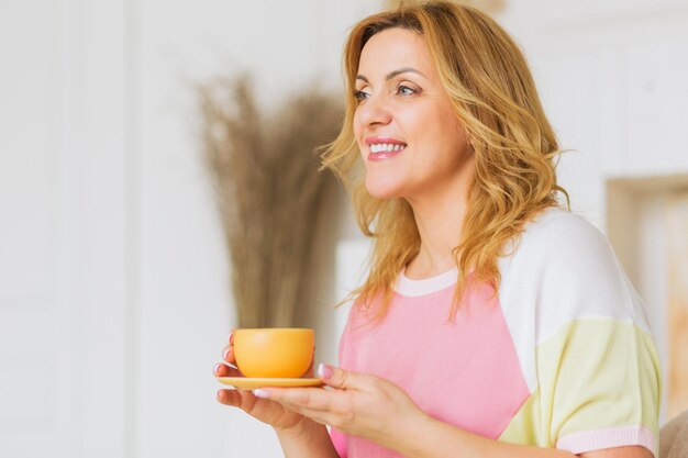 Uma linda mulher madura em um suéter rosa e calça amarela posa para a câmera com uma xícara de chá. Conceito de vida feliz da mulher após 40 anos