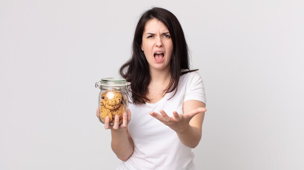 Uma linda mulher parecendo zangada, irritada e frustrada, segurando uma garrafa de vidro de biscoitos