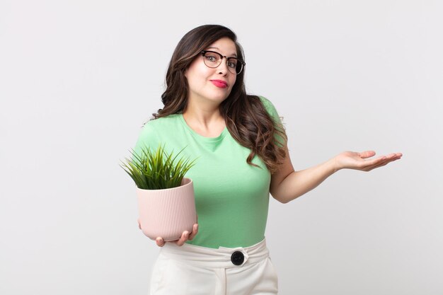 Uma linda mulher se sentindo perplexa e confusa, duvidando e segurando uma planta decorativa