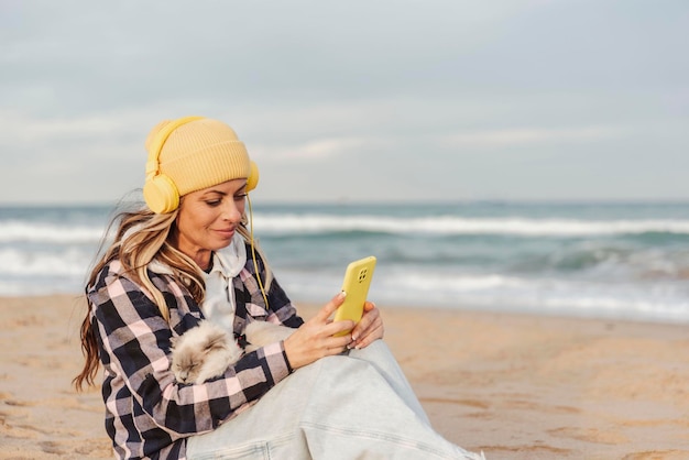 Uma linda mulher sentada na praia com seu gato usando um telefone inteligente e ouvindo música com fones de ouvido