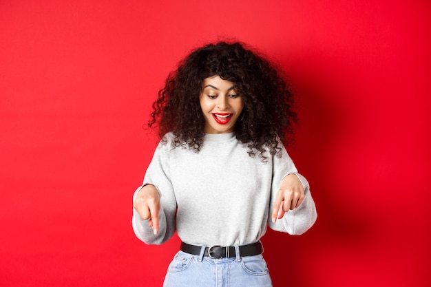 Uma linda mulher sonhadora com cabelo encaracolado, apontando e olhando para baixo animada, conferindo a promo, em pé contra um fundo vermelho.