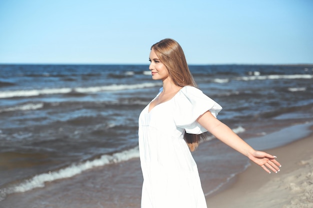 Uma linda mulher sorridente e feliz está na praia do oceano com um vestido branco de verão, braços abertos