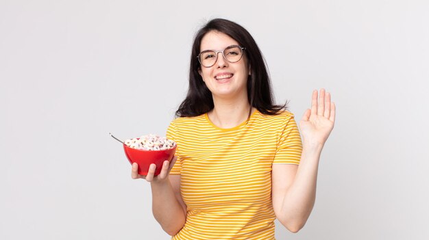 Uma linda mulher sorrindo feliz, acenando com a mão, dando as boas-vindas, cumprimentando você e segurando uma tigela de café da manhã