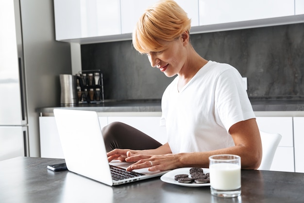 Uma linda mulher usando um laptop enquanto está sentado à mesa da cozinha, bebendo leite em um copo, comendo biscoitos