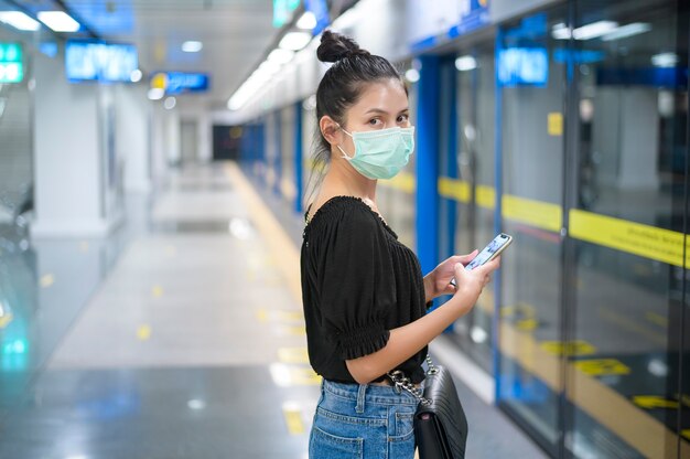 Uma linda mulher usando uma máscara cirúrgica usando o telefone enquanto espera um trem no metrô, tecnologia e conceito covid19