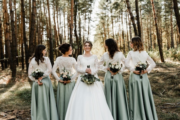 Uma linda noiva de aparência europeia segura um buquê de casamento nas mãos e olha para as damas de honra que seguram buquês de flores nas costas