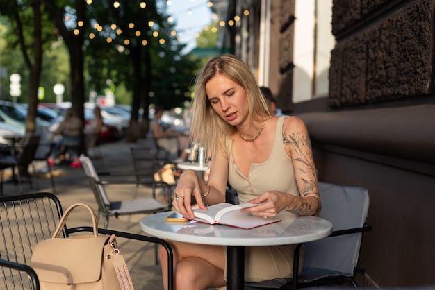 Uma loira com uma tatuagem no braço está sentada em um café de verão e fala emocionada ao telefone