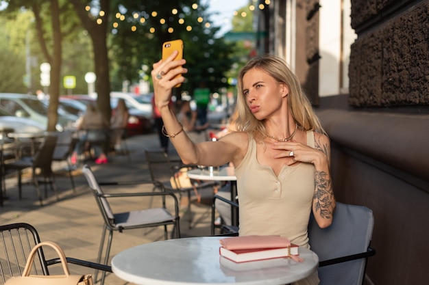 Uma loira com uma tatuagem no braço está sentada em um café de verão e fala emocionada ao telefone
