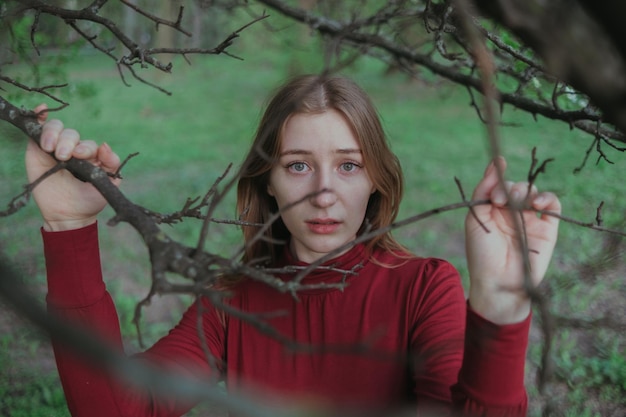 Uma loira de vermelho olha para a câmera através dos galhos de uma árvore