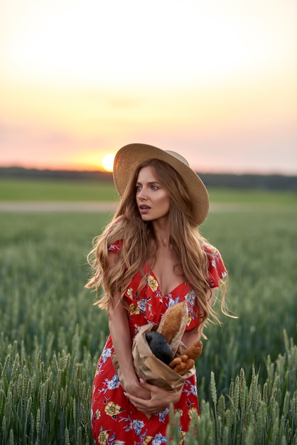 Uma loira de vestido vermelho e chapéu de palha com pão francês nas mãos em um campo de trigo ao pôr do sol