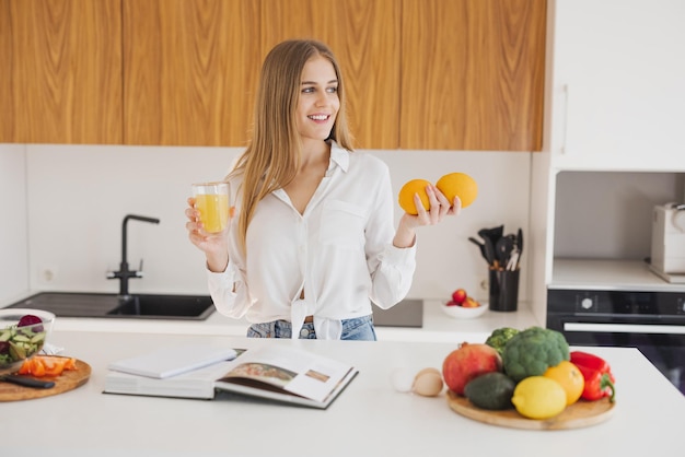 Uma loira feliz está olhando para um livro de receitas e bebendo suco na cozinha