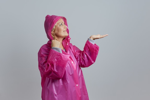 Uma loira madura e sorridente em uma capa de chuva magenta posando com um capuz, estendendo a mão para as gotas de chuva. Isolado em fundo cinza.