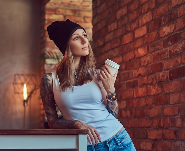 Uma loira sexy tatuada em uma t-shirt e um chapéu com uma xícara de café em uma sala com interior loft.